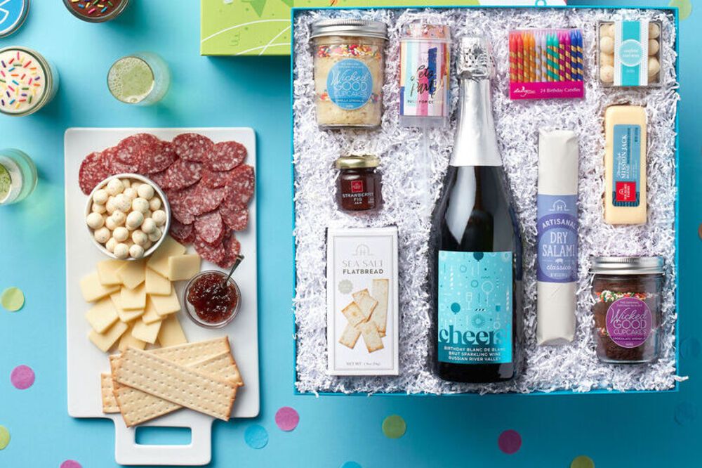 A blue gift box filled with a variety of food items, including crackers, cheese, a bottle of wine, and artisan dry salami. The box is on a table, and there are candles on the table.