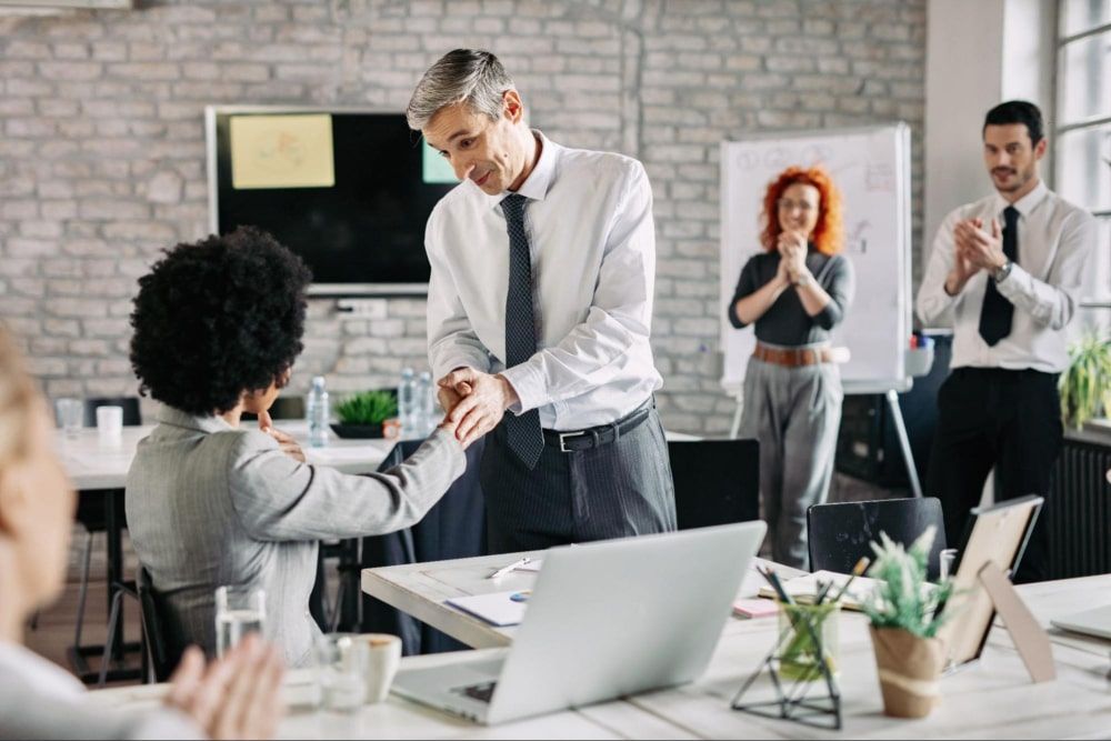 Happy businessman and his female colleague shaking hands in the office after a successful agreement, while other coworkers applaud them