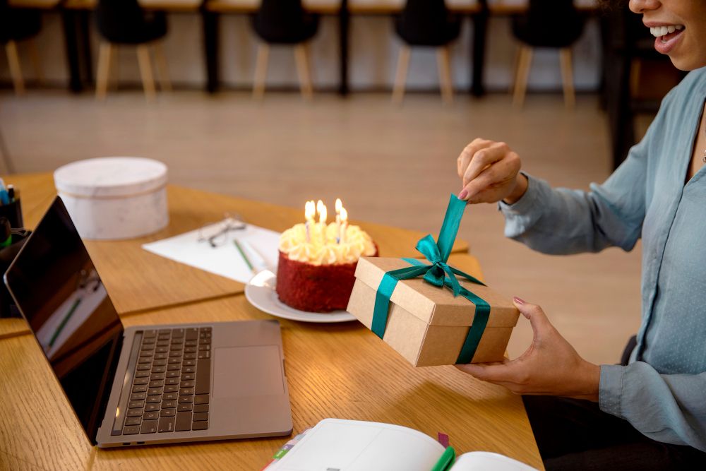 Woman sitting at a table with a laptop and a cake while opening a present.