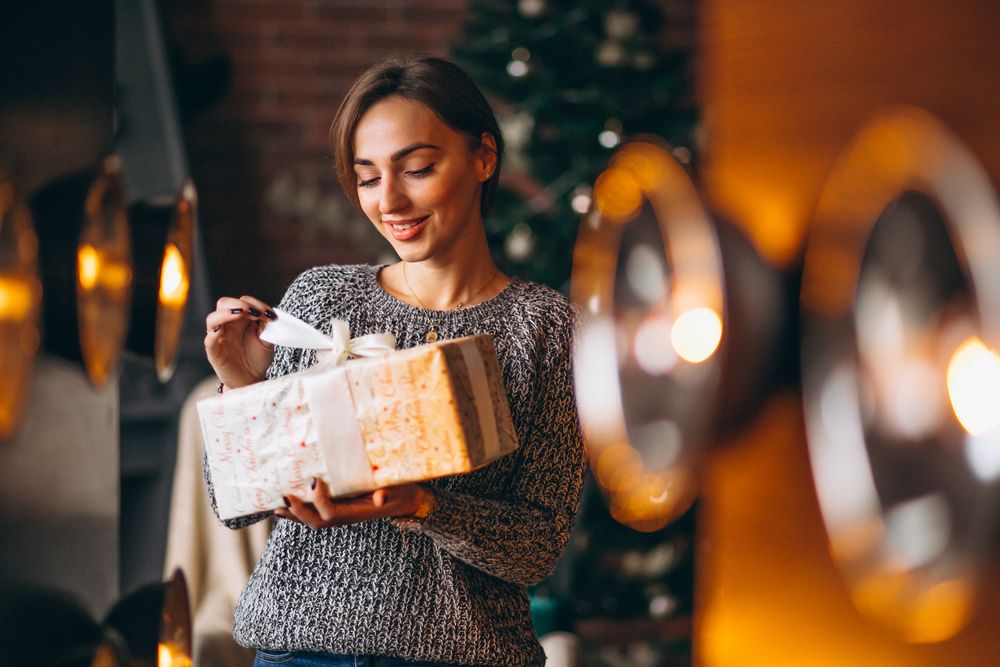 Woman with a present by a Christmas tree harmonizes with the idea of choosing client gifts for Christmas, encapsulating the spirit of thoughtful and opulent giving.